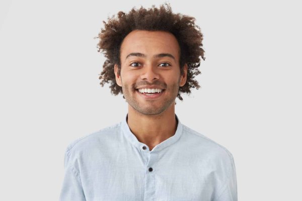 Cheerful positive glad mixed race male has broad smile, rejoices promotion at work, recieves congratulations from colleagues, dressed in white shirt, stands alone in studio, has satisfied expression
